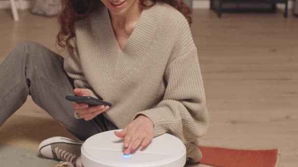 若いブルネット白人女性は 居心地の良いリビングルームで床に座って 新しいロボット真空を設定しながら 彼女の携帯電話を使用しています — ストック動画