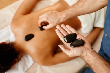 Genç bir adamın rahatlatıcı SPA oturumunda genç bir bayanın sırtına taş koyduğu yakın plan.