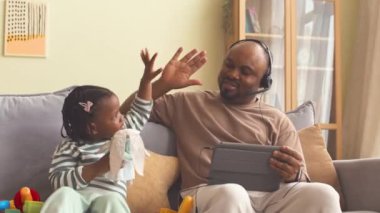 Mutlu Afro-Amerikalı adam evinden dijital tablet üzerinde çalışırken küçük tatlı kızıyla oynuyor.