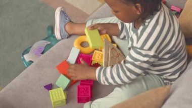 3 yaşındaki Afro-Amerikalı güzel, çizgili, uzun kollu tişörtlü bir kızın oturma odasındaki koltukta renkli oyuncak bloklarıyla oynaması.