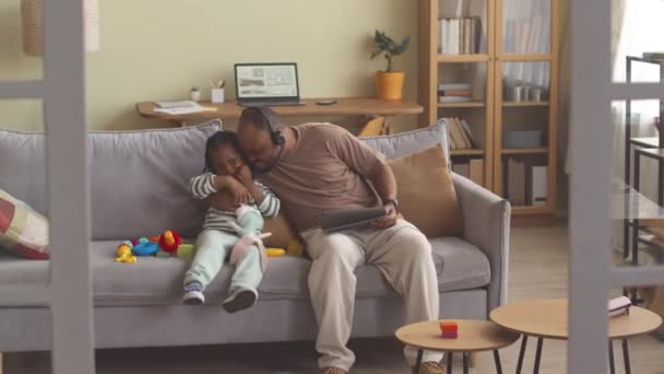 居心地の悪いリビングルームでソファーに座っている間 彼の3歳の娘を抱きしめて家から働く黒人の男 — ストック動画
