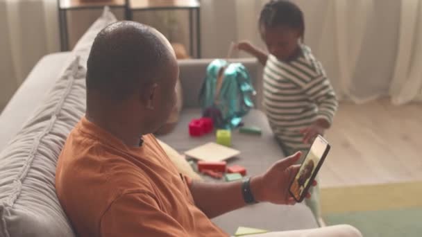 Афроамериканец Видео Чата Смартфоне Время Симпатичная Дочка Играет Игрушками Диване — стоковое видео