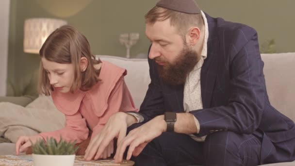 当坐在客厅咖啡桌旁的沙发上的时候 犹太父亲穿着Kippah和小学女生一起解开拼图 — 图库视频影像