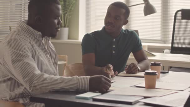 Dwóch Młodych Afroamerykanów Omawia Dokumenty Biznesowe Siedząc Razem Przy Biurku — Wideo stockowe