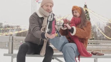 Genç beyaz adam, kış tatillerinde buz patenlerinde dışarıda buluşurken kız arkadaşının buz patenlerine ayakkabı bağcığı bağlamasına yardım ediyor.