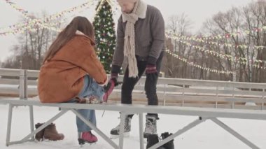 Genç beyaz adam, kış tatilinde buz patenlerinde buz pateni pistinde romantik bir randevu yaşarken kız arkadaşına ayakkabı bağcıklarını bağlamasında yardım ediyor.