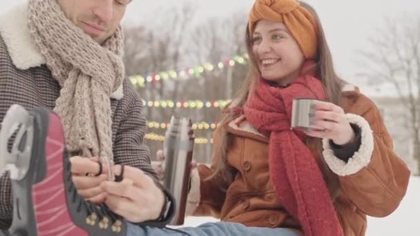 美しい若い白人女性は 彼女のボーイフレンドが氷のスケートで靴ひもを結んで 冬の休日にアイスリンクで屋外でロマンチックな日付を持っている間 熱いお茶を楽しんでいます — ストック動画