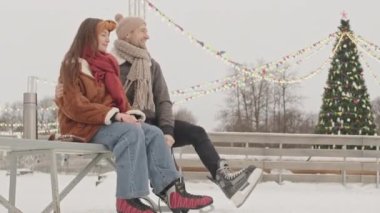 Sevecen genç çift buz patenine hazırlanırken eğleniyor, arka planda çelenkleri yanan uzun Noel ağacıyla dışarıda bankta oturuyor.