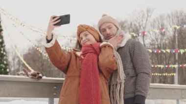 Birbirine aşık genç bir çift, arka planda güzel dekore edilmiş uzun Noel ağacıyla buz pateni pistinde beklerken akıllı telefondan selfie çekiyor.