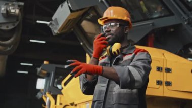 Siyah erkek traktör fabrika çalışanı, iş yerinde iş arkadaşlarını telsiz kullanarak koordine ediyor.