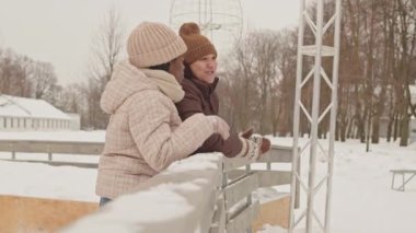 Genç, çok ırklı lezbiyen çift, kış günü buz pateni pistinde dikilirken tahta korkuluklara yaslanıp sohbet ediyorlar.