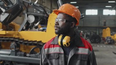 Kasketli ve güvenlik gözlüklü kendinden emin siyah adam fabrikada yürüyor traktör, buldozer ve diğer makine ekipmanları üretiyor.