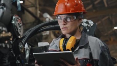 Ciddi genç bayan mühendis elinde dijital tabletle traktör fabrikasında modern makine ekipmanlarını inceliyor.