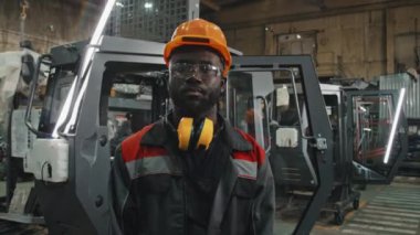 Modern makine ekipmanlarına karşı duran kameraya bakan genç Afrikalı Amerikalı erkek traktör işçisinin orta boy portresi.
