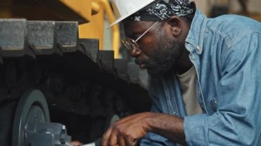 Fabrikadaki traktör makinesini tamir ederken sürgülü anahtar kullanan kot gömlekli ve beyaz başlıklı profesyonel Afro-Amerikan tamircinin yavaşlaması