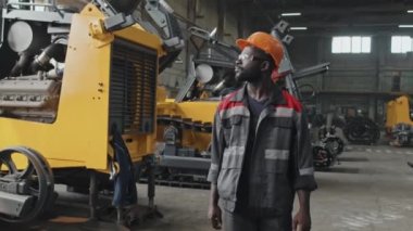 Profesyonel Afro-Amerikan erkek işçi, güvenlik gözlükleri ve baret takıyor traktör üretim tesisinde yürüyor ve araç örneklerine bakıyor.