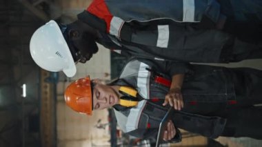 Elinde dijital tabletle kadın traktör yöneticisinin dikey görüntüsü. Makine ekipmanlarının yanında duran genç siyahi adamla konuşuyor.