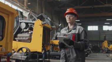 Güvenlik gözlüklü genç kadın mühendisin orta boy portresi, başlık ve kapak dijital tablet taşıyor ve traktör fabrikasında kameraya poz veriyor.