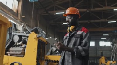 Genç siyahi erkek mühendisin, fabrikadaki traktörleri ve diğer makineleri incelerken tablet bilgisayar kullanırken orta boy görüntüsü.