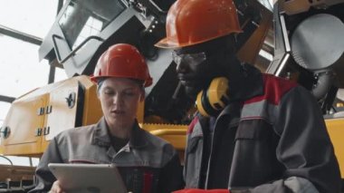 Tulumlu ve şapkalı birkaç çok ırklı mühendis tablet bilgisayar üzerinde çalışıyor ve traktör üretim tesisini incelerken konuşuyorlar.
