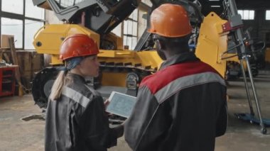 Endüstriyel fabrikada birlikte çalışırken dijital tablette traktör makine şemasını tartışan iki çoklu etnik mühendisin arka görünümü