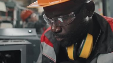 Afrika kökenli Amerikalı güvenlik gözlükleri ve başlık takan mühendislerin endüstriyel fabrikada bayan meslektaşlarıyla çalışırken tablet bilgisayar kullanmaları.