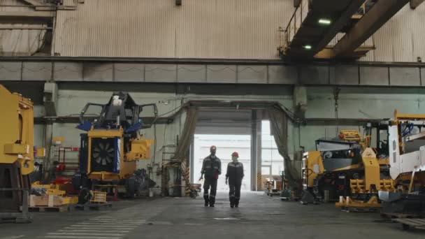 两个不同的拖拉机制造厂工人 穿着工作服 头戴硬礼帽 轮流在车间边走边聊天 — 图库视频影像