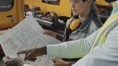 Sert şapka ve asit yeşili yelek giyen profesyonel siyah erkek mühendis kadın traktör fabrika işçisine teknik entrika gösteriyor.