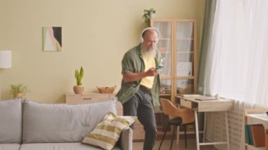 Telsiz kulaklıklı mutlu sakallı adam evde tek başına vakit geçirirken akıllı telefon dinleyip dans ediyor.