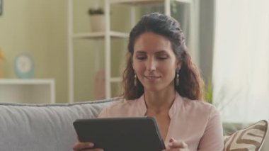 Çekici esmer kadın, modern oturma odasındaki koltukta rahatça oturup dijital tablet kullanıyor.