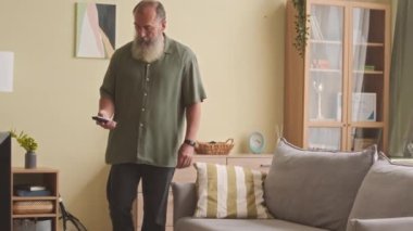 Uzun gri sakallı, sıkıcı beyaz bir adam TV 'de kanal değiştiriyor, öğleden sonra oturma odasında kanepede oturuyor.