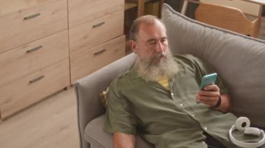 Sakallı, yaşlı, beyaz bir adam evinde koltukta dinleniyor. Akıllı telefondan kayıyor ve TV kanallarını değiştiriyor.