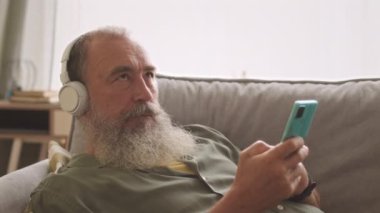 Modern son sınıf öğrencisi evdeki koltukta akıllı telefonuyla dinlenirken kablosuz kulaklıkla müzik dinliyor.