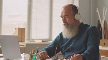 Başarılı, kablosuz kulaklıklı, yaşlı, beyaz, başarılı bir iş adamı. Bilgisayarlı bir ekiple görüntülü konuşma yapıyor. Modern ofiste masa başında oturuyor.