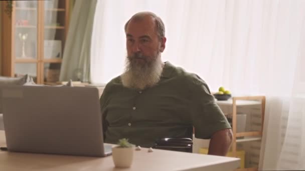 坐在轮椅上 头戴扬声器耳机的白种人老人在办公室的笔记本电脑上工作 — 图库视频影像