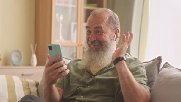 白胡子老人 头戴无线耳塞 在智能手机上与家人聊天 白天坐在客厅沙发上 — 图库视频影像