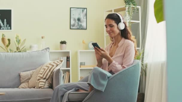 在舒适的公寓里 快乐的白人年轻女人喜欢用无线耳机听她最喜欢的智能手机播放音乐 — 图库视频影像