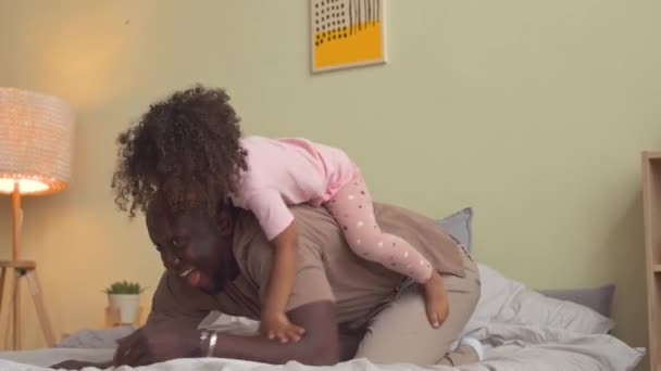 在可爱的粉色睡衣中嬉笑着的非洲裔美国小女孩 一边骑在她可爱的父亲的背上 一边在床上玩耍 — 图库视频影像