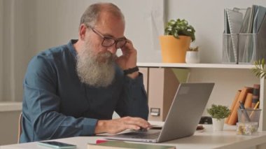 Ofiste dizüstü bilgisayarla çalışırken sigara içen kendinden emin, beyaz sakallı, yaşlı bir iş adamı.