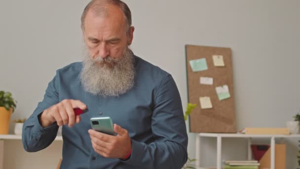 现代白人大胡子男子在智能手机上打滚 一边微笑 一边在工作场所吸烟的中景照片 — 图库视频影像
