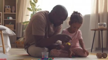 Afrikalı Amerikalı bir adam ve küçük kızı rahat bir dairede otururken renkli mankenlik killeriyle oynuyorlar.