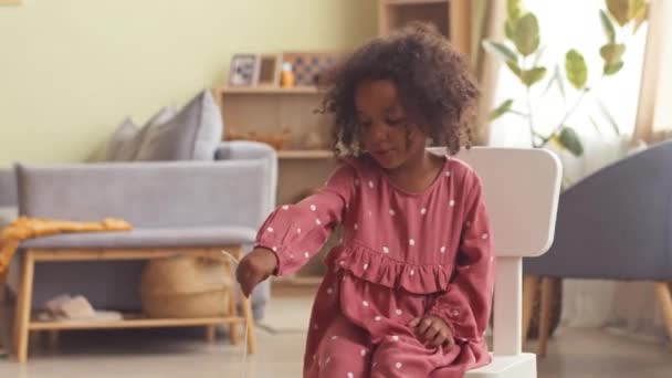 かわいいアフリカ系アメリカ人の幼い少女の肖像画は 居心地の良いリビングルームに子供の椅子に座っている間 カメラを見ているかわいいポルカのドットドレス — ストック動画