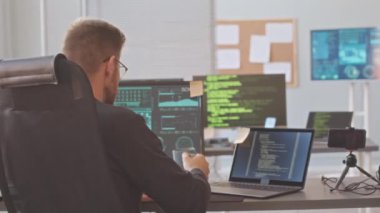 Gözlüklü beyaz erkek programcının orta boy portresi kameraya dönüyor ve gülümsemeye başlıyor, program kodları ve bilgisayar ve dizüstü bilgisayar şemalarıyla ofis masasında oturuyor.