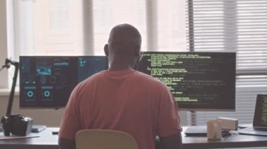 Ofis masasında iki bilgisayar monitörü ve dizüstü bilgisayar başında oturmuş program kodları yazan Afrikalı Amerikalı yazılım mühendisinin arka görüntüsü