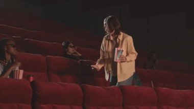 Beyaz kız sinema salonundaki yerleri gezerken akıllı telefonuna meşale takıyor, oturuyor ve 3D gözlükler takıyor.