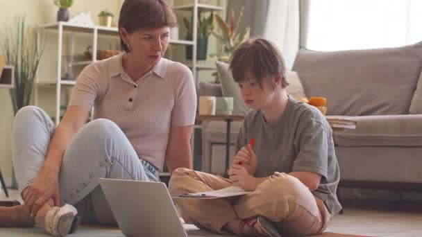 14岁的患有下行综合症的女孩通过笔记本电脑在线学习 她坐在客厅的地板上 由她有爱心的母亲帮助和支持她 — 图库视频影像
