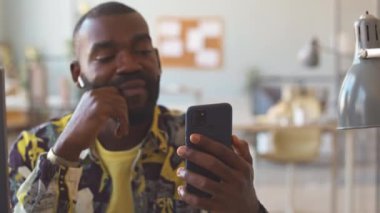 Renkli baskılı gömlek videosundaki genç siyahi adam iş yerindeki iş yerinde oturan kablosuz kulaklıklardan iş arkadaşıyla birlikte akıllı telefondan arıyor.