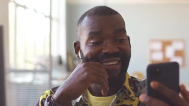 Kablosuz kulaklıklı Afrikalı Amerikalı bir adam ofisteki meslektaşıyla akıllı telefondan görüntülü sohbet ediyor.