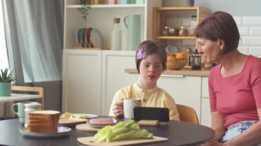 Down sendromlu 14 yaşında bir kız annesiyle mutfak masasında oturmuş kahvaltıda ya da öğle yemeğinde akıllı telefondan video seyrediyor.