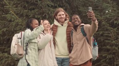 Bir grup farklı genç arkadaş, baharda çimen ağaçlarıyla parkta yürürken çekilmiş videolarını çekerken ve akıllı telefon kamerasına gülümsüyor ve el sallıyorlardı.
